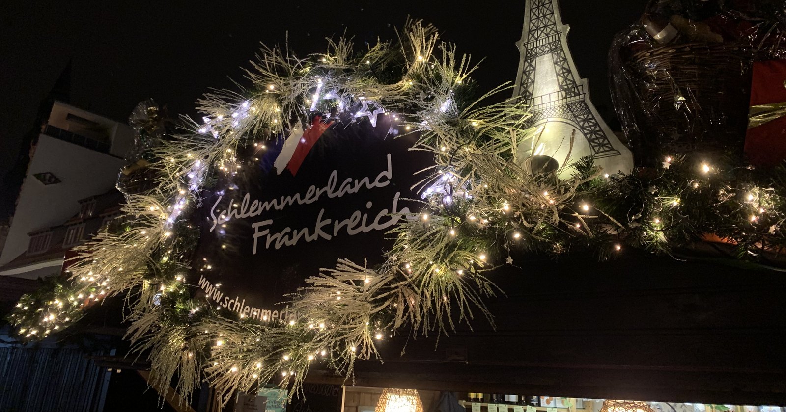 Beleuchteter Tannenkranz mit Eiffelturm-Motiv über dem Stand bei Nacht, dahinter Lichterketten